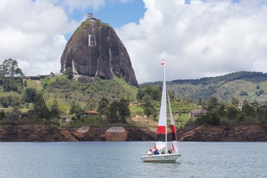 Ein Segelboot mit mehreren Personen gleitet über den ruhigen Stausee, während der markante El Peñol im Hintergrund thront.