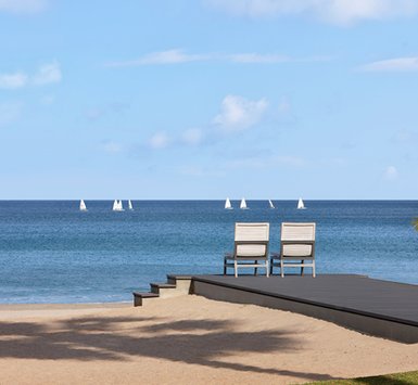 Zwei Liegestühle stehen auf einer Holzterrasse am Strand, mit Blick auf das ruhige, blaue Wasser und Segelboote in der Ferne.