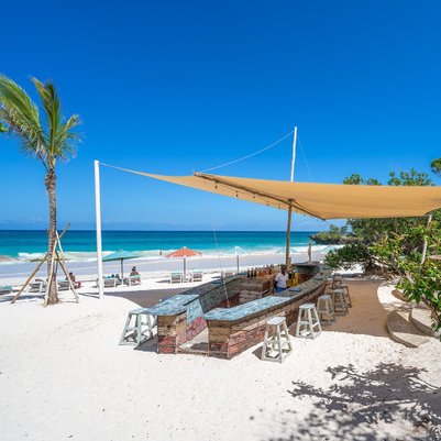 Ein einladender Strand mit einem Holzbar, umgeben von Palmen und Sonnenschirmen, unter einem strahlend blauen Himmel.