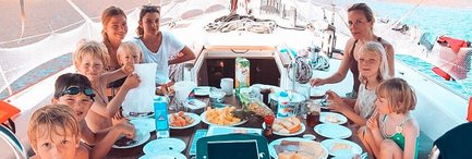 Eine fröhliche Gruppe von Kindern und Erwachsenen genießt ein gemeinsames Frühstück auf einem Segelboot, umgeben von Wasser.