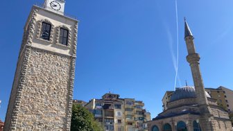 Eine steinerne Uhrturm mit einem Ziffernblatt und ein Minarett stehen unter einem klaren blauen Himmel in Korça.