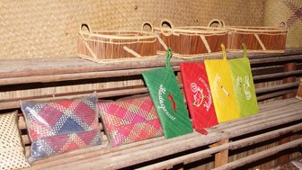 Bunte, handgefertigte Taschen aus Madagascar liegen auf einem Holzregal, umgeben von geflochtenen Körben im Hintergrund.
