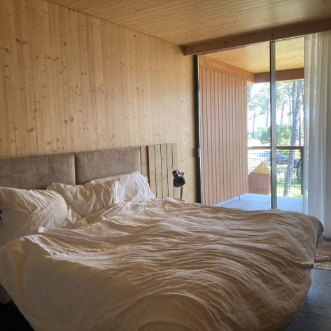Ein helles Schlafzimmer mit einem großen Bett, umgeben von Holzverkleidung und einem Blick auf die Natur durch große Fenster.