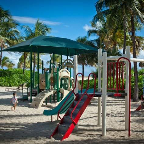 Ein Kind in einem bunten Badeanzug läuft fröhlich auf einem Spielplatz am Strand unter Palmen.
