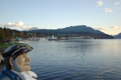 Eine Statue eines alten Seemanns blickt auf das ruhige Wasser, während die Küstenstadt im Hintergrund sichtbar ist.
