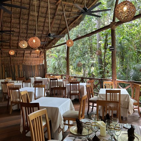 Ein stilvolles Restaurant mit Holzmöbeln und eleganten Tischen, umgeben von üppiger tropischer Vegetation.