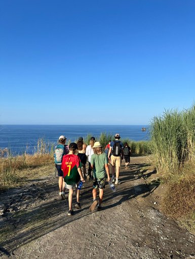 Eine Gruppe von Wanderern geht einen schmalen Pfad entlang, umgeben von hohem Gras und mit Blick auf das blaue Meer.
