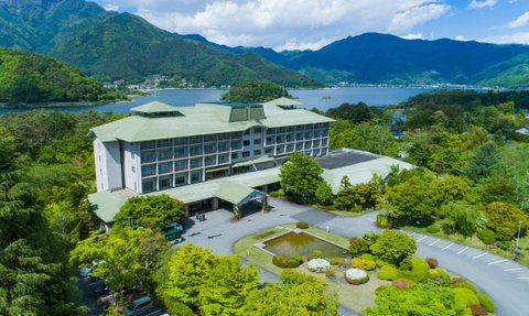 Ein modernes Hotel mit grünem Dach, umgeben von üppiger Natur und Blick auf einen ruhigen See und die Berge im Hintergrund.