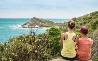 Zwei Personen sitzen auf einem Felsen und blicken auf das türkisfarbene Wasser und die Küste in der Ferne.