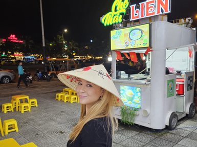 Eine lächelnde Frau trägt einen traditionellen vietnamesischen Hut und steht vor einem Straßenstand mit bunten Stühlen.