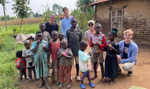 Eine Gruppe von Kindern steht fröhlich im Freien, umgeben von Natur und einem einfachen, rustikalen Hintergrund.