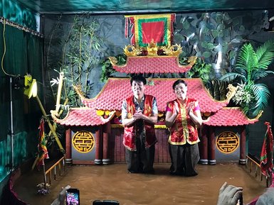 Zwei Puppenspieler stehen in knalligen Kostümen vor einer traditionellen Kulisse, umgeben von Wasser und Pflanzen.