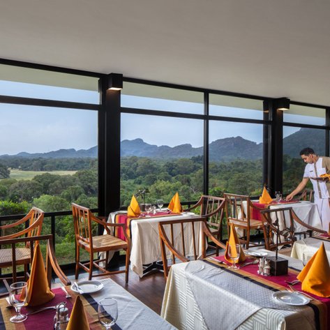 Ein elegantes Restaurant mit Blick auf eine grüne Landschaft und Berge, während ein Kellner den Tisch deckt.