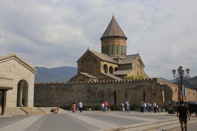 Eine beeindruckende Kirche mit einer markanten Kuppel steht vor einer alten Stadtmauer, umgeben von Besuchern und Bergen.