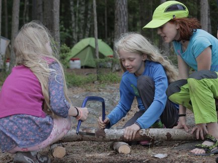 Drei Kinder arbeiten zusammen, um einen Baumstamm im Wald mit einer Handsäge zu zersägen, während Zelte im Hintergrund stehen.