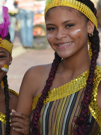 Zwei fröhliche Mädchen in bunten Kostümen lächeln in die Kamera, umgeben von einer lebhaften Menschenmenge.