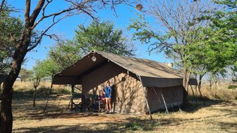Ein Zeltcamp in der Natur, umgeben von Bäumen und Gras, mit einer Familie, die entspannt am Tisch sitzt.