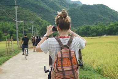 Eine Frau mit einem bunten Rucksack fotografiert die Landschaft in einem Reisfeld, umgeben von grünen Bergen.