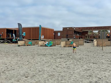 Ein Junge läuft barfuß über den Sandstrand, während im Hintergrund eine Strandbar und Liegestühle zu sehen sind.