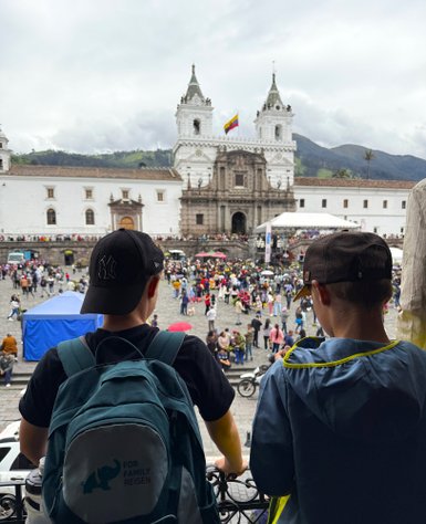 Zwei Jungen stehen an einem Balkon und beobachten eine lebhafte Menschenmenge auf einem Platz vor einer beeindruckenden Kirche.