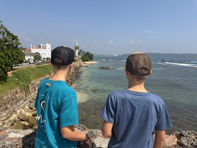 Zwei Jungen stehen am Ufer und blicken auf das Meer, während ein Leuchtturm in der Ferne sichtbar ist.