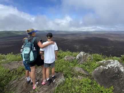 Eine Mutter steht mit zwei Kindern an einem Aussichtspunkt und blickt auf eine weite, vulkanische Landschaft.