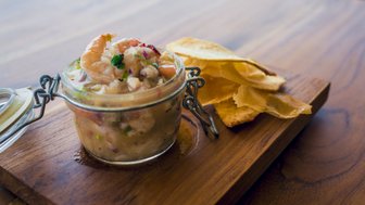 Ein Glas mit frischem Ceviche, garniert mit Garnelen und Kräutern, steht auf einem Holzbrett neben knusprigen Tortilla-Chips.