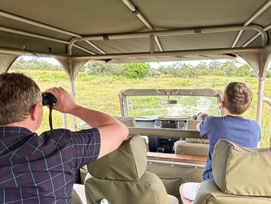 Zwei Personen sitzen in einem Geländewagen und beobachten die Natur, während ein Elefant im Hintergrund sichtbar ist.