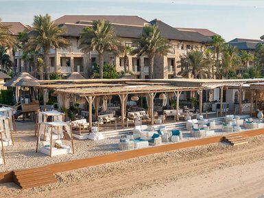 Ein luxuriöser Strandbereich mit gemütlichen Liegen und einem stilvollen Restaurant im Hintergrund, umgeben von Palmen.