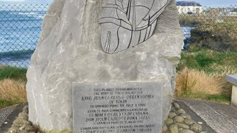 Ein großer, grauer Stein mit einer eingravierten Schiffsdarstellung steht an der Küste, umgeben von sanften Wellen und Gras.