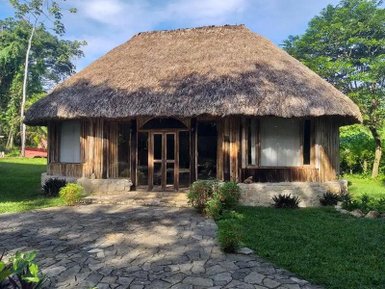 Ein traditionelles, strohgedecktes Haus aus Holz steht in einer grünen Umgebung mit einem gepflasterten Weg davor.
