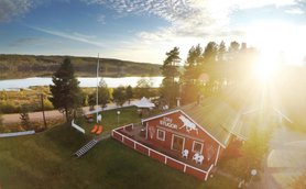 Ein rotes Ferienhaus mit einem weißen Pferdemotiv steht in einer malerischen Landschaft am Wasser, umgeben von Bäumen.