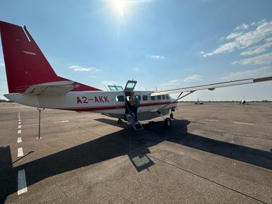 Ein kleines Flugzeug mit rotem Heck steht auf dem Rollfeld, während Passagiere einsteigen und die Sonne scheint.