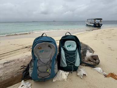 Zwei Rucksäcke stehen im Sand, umgeben von Muscheln, während im Hintergrund ein Boot sanft im Wasser schaukelt.