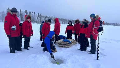 Eine Gruppe von Menschen in roten Jacken steht um ein Loch im Eis, während einer von ihnen mit einem Löffel arbeitet.