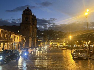 Die Plaza de Armas in Cusco erstrahlt in warmem Licht, während Autos und Motorräder die gepflasterte Straße entlangfahren.