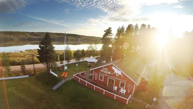 Ein rotes Ferienhaus mit einem weißen Pferdemotiv steht in einer malerischen Landschaft am Wasser, umgeben von Bäumen.