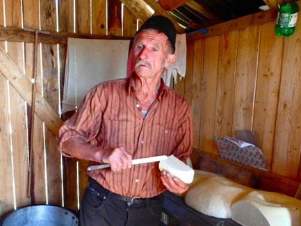 Ein älterer Mann in einem Holzraum hält ein Stück Käse und schaut überrascht, während er mit einem Messer arbeitet.