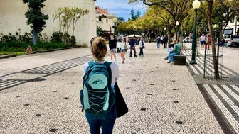 Eine Person mit einem Rucksack geht auf einem gepflasterten Weg, umgeben von Bäumen und anderen Spaziergängern.