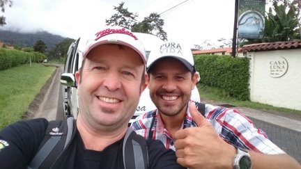Zwei Männer posieren fröhlich für ein Selfie, während im Hintergrund eine grüne Landschaft und ein Schild mit 'CASA LUNA' sichtbar sind.