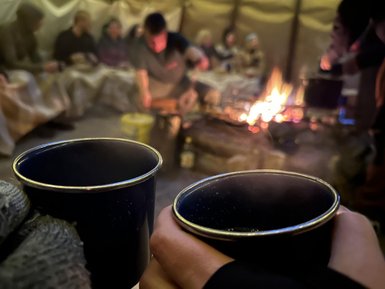 Zwei dampfende Tassen werden in der Nähe eines Lagerfeuers gehalten, während Menschen im Hintergrund entspannt sitzen.