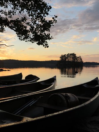 Am Ufer eines ruhigen Sees liegen mehrere Kanus, während die Sonne hinter den Bäumen untergeht und den Himmel erleuchtet.