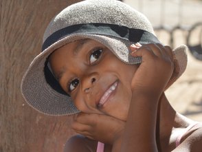 Ein fröhliches Kind mit einem grauen Hut lächelt und stützt das Gesicht auf die Hände, während es vor einem Baum steht.