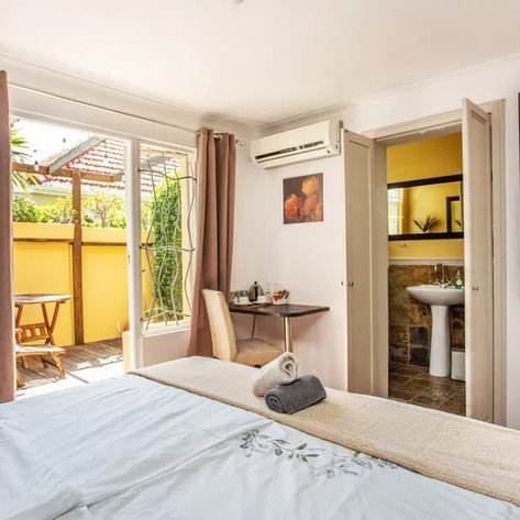 Ein helles Zimmer mit einem großen Bett, einem Tisch und einem Stuhl, das auf eine sonnige Terrasse mit Pflanzen blickt.