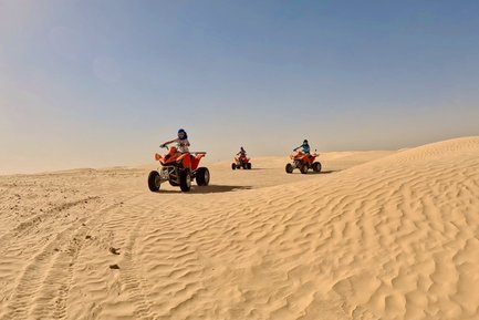 Drei Personen fahren auf Quads über die sanften Sanddünen einer Wüstenlandschaft unter klarem Himmel.