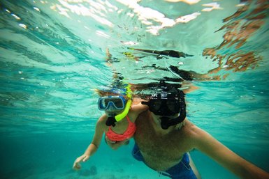 Ein Erwachsener und ein Kind schnorcheln gemeinsam unter Wasser, umgeben von klarem, blauem Wasser und Lichtreflexionen.
