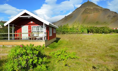 Ein rotes Ferienhaus mit einer Veranda steht vor einem majestätischen Berg, umgeben von grünen Wiesen und blühenden Pflanzen.