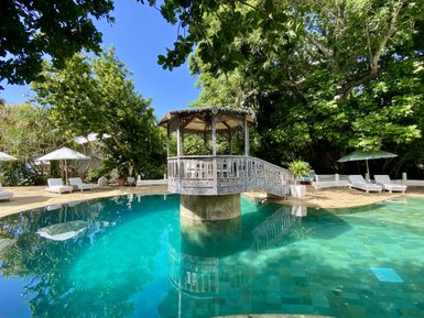 Ein ruhiger Poolbereich mit klarem Wasser, umgeben von üppigem Grün und einem charmanten Pavillon im Hintergrund.