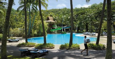 Umgeben von Palmen liegt ein glitzernder Pool, während ein Servicekraft eilends Handtücher bringt und das Resort belebt.