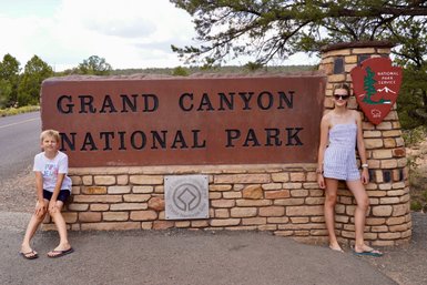 Ein Junge und ein Mädchen posieren lächelnd vor dem Eingangsschild des Grand Canyon Nationalparks.
